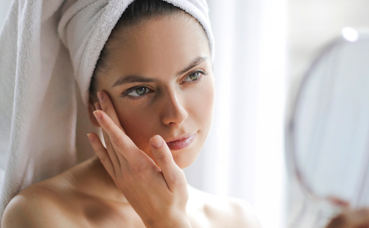 a woman with a towel wrapped around her head, looking and touching at her skin in the mirror with a content expression, indicating a post-bath self-care routine