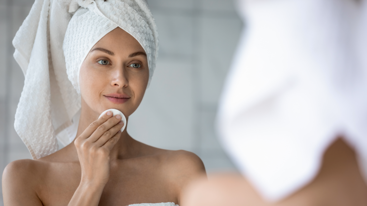 fresh-looking woman with a towel on her head, cleaning her chin in the mirror with a puff after a shower