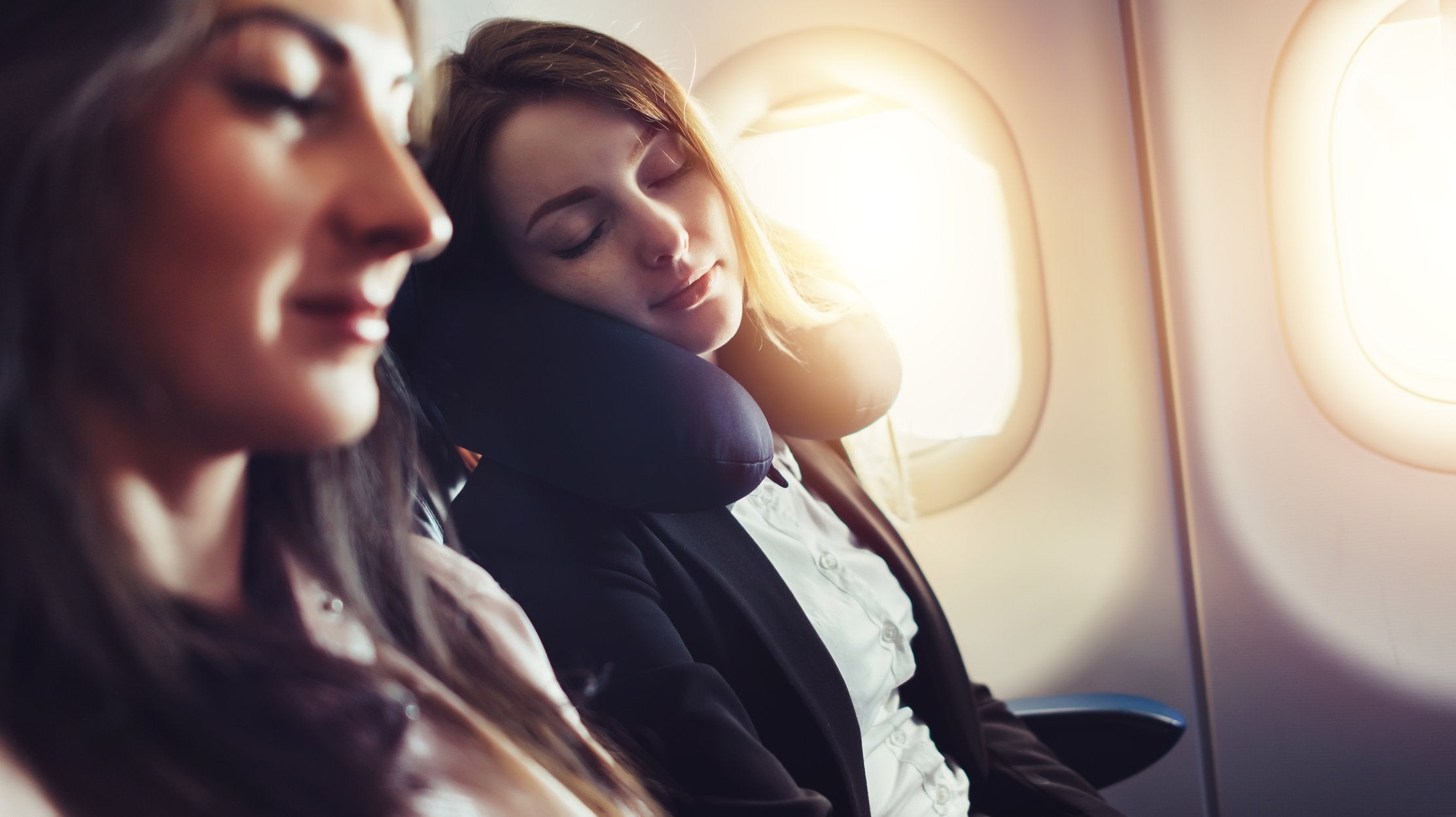 two women sitting inside a plane, with one sleeping in the back and the other slightly blurry in the front, both wearing relaxed facial expressions