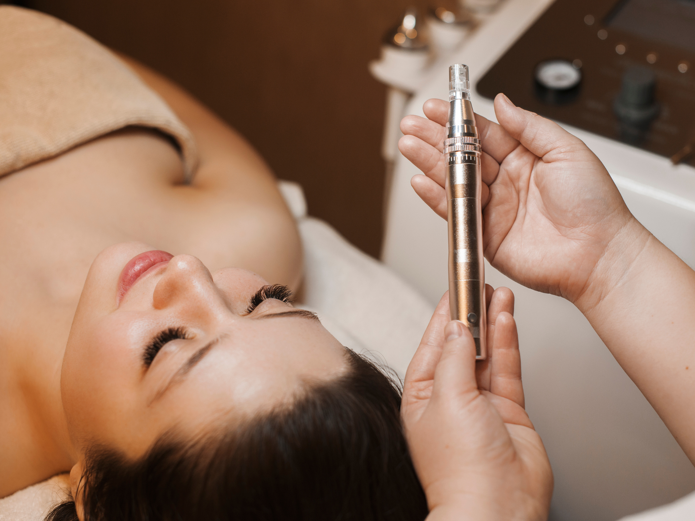 top view of cosmetologist's hands holding a Dermapen for microneedling procedure on a woman's face, who is leaning on a bed with closed eyes