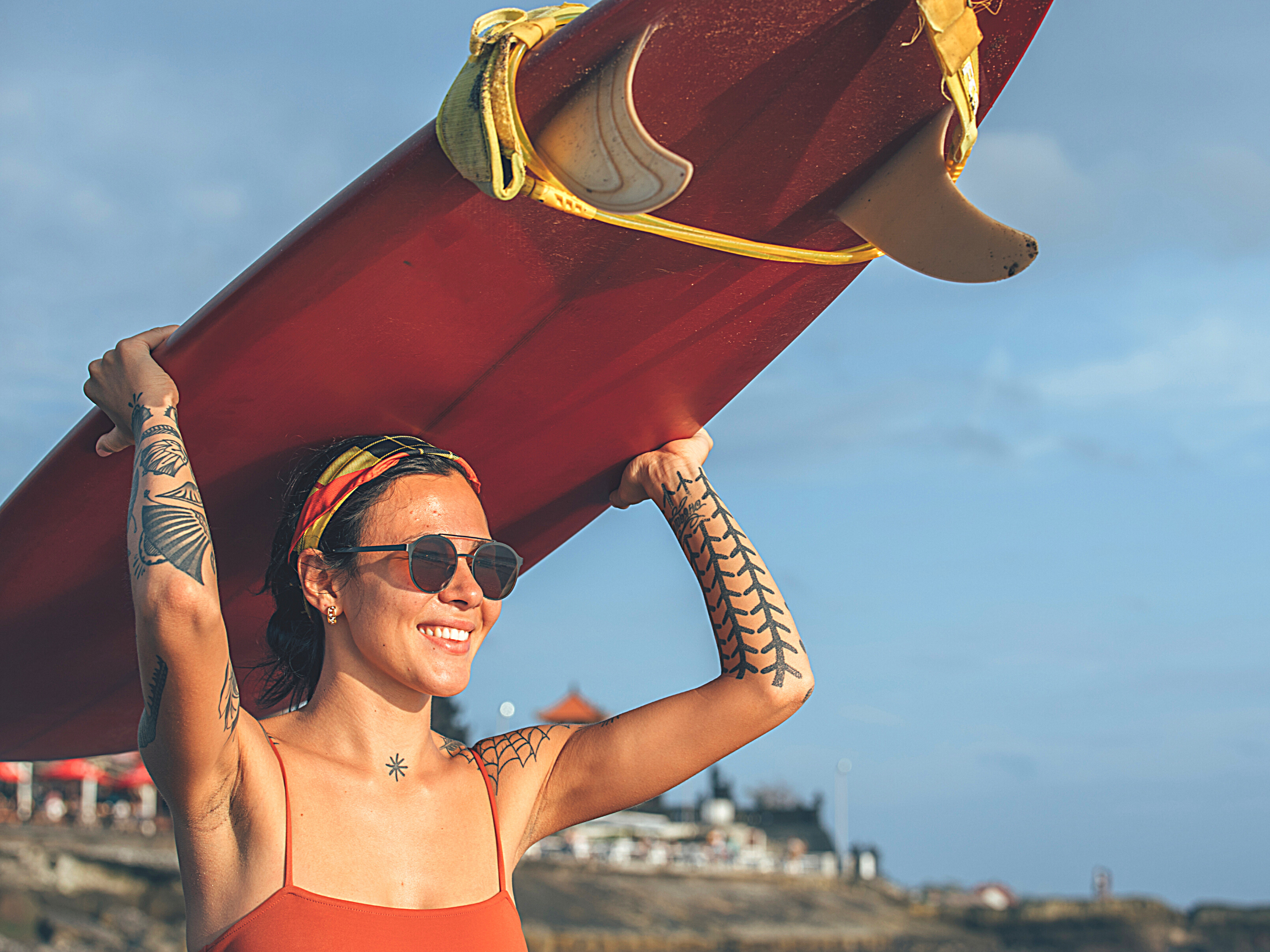 a smiling woman with tattoos wearing sunscreen with a surfboard on her head walking towards the beach for water surfing