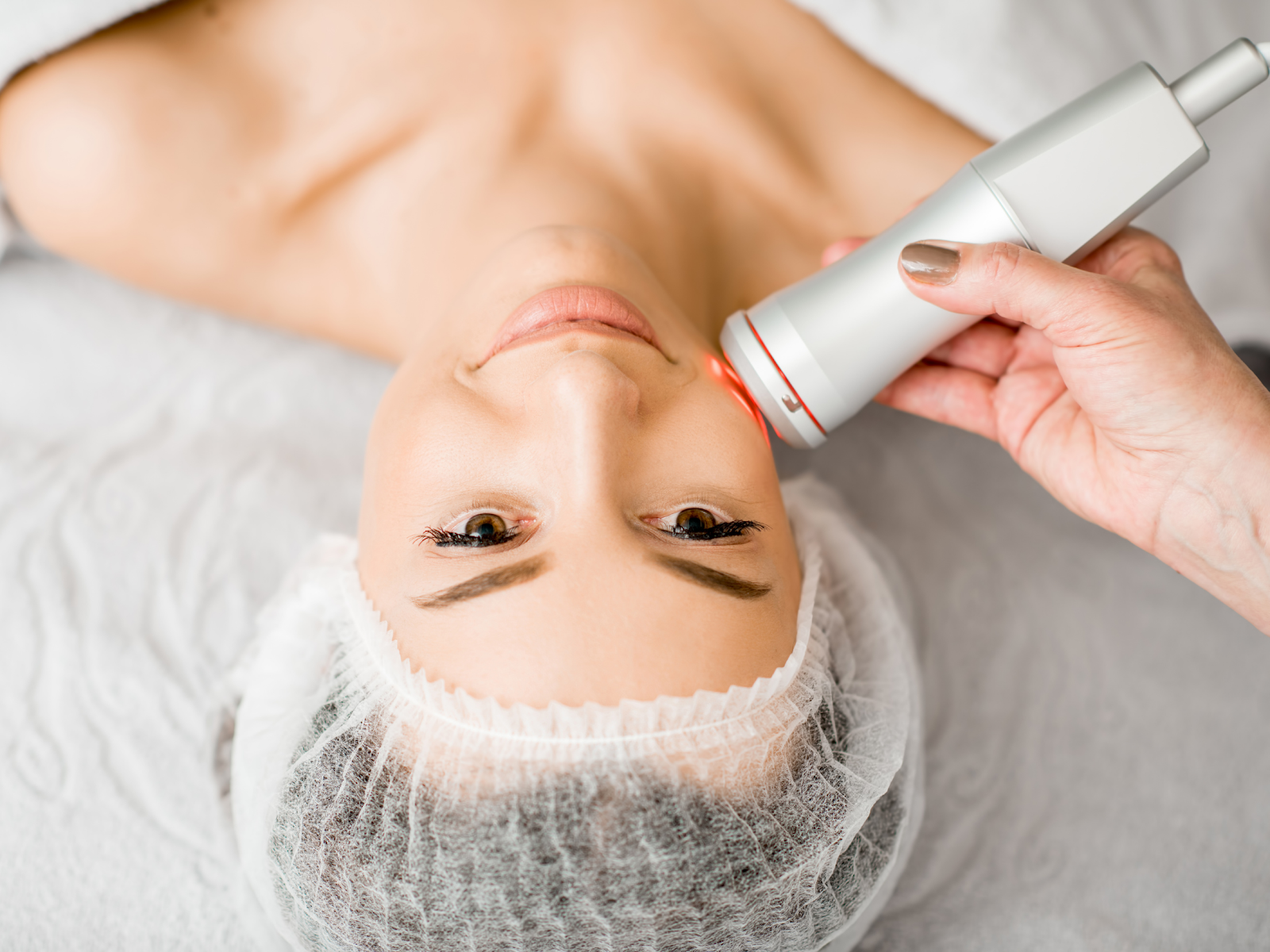 a woman laying on a treatment bed at a cosmetology clinic, receiving a skincare massage with an electrical tool, indicating a professional skincare treatment